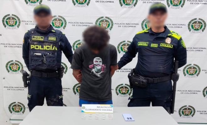 Dos funcionarios policiales con un ciudadano capturado frente a pendón institucional frente a ellos mesa sobre la cual esta el material incautado