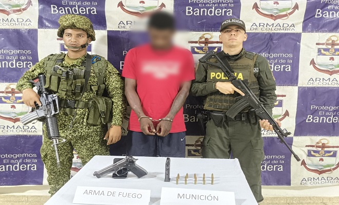Un integrante de la Armada Nacional, una persona capturada y un uniformado de la Policía Nacional posan junto a una mesa donde se observa un arma de fuego tipo pistola, un proveedor y varios cartuchos de munición incautados