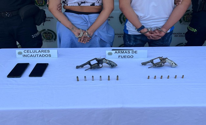 Elementos incautados, entre ellos dos armas de fuego y dos celulares, de fondo dos personas capturadas y dos funcionarios de la Policía Nacional