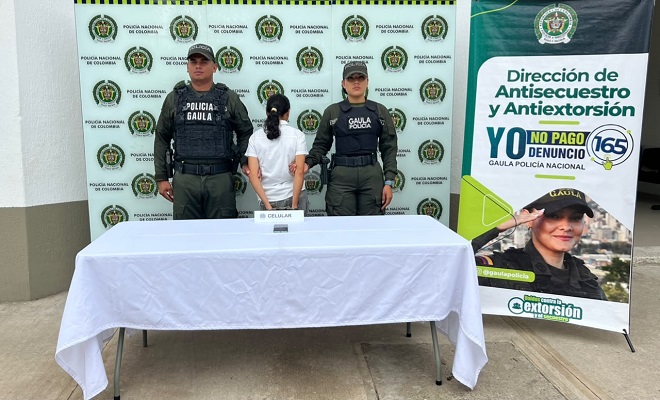Un celular incautado sobre una mesa con mantel blanco, una persona de sexo femenino capturada por el delito de extorsión, acompañada de dos uniformados del GAULA de la Policía Nacional brindando seguridad.