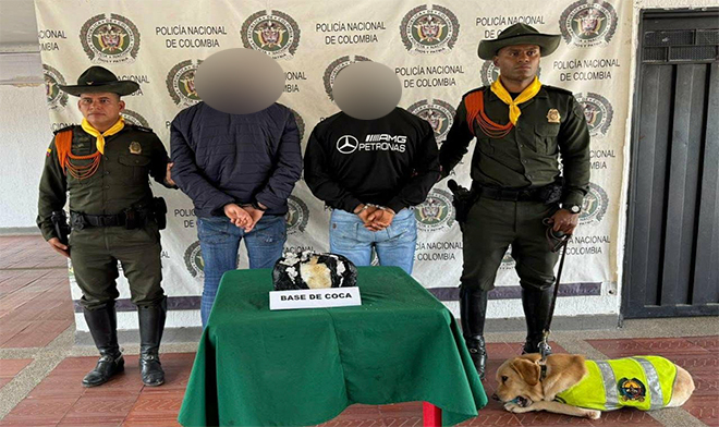 Dos policías,  dos personas capturadas , una bolsa de estupefaciente encima de una mesa y un perro.