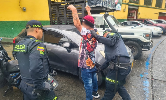 Dos uniformados de la Policía están realizando un registro   a un hombre que tiene los brazos levantados contra un vehículo gris