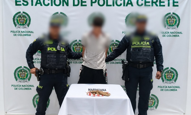 Dos funcionarios policiales con un ciudadano capturado frente a pendón institucional frente a una mesa sobre la cual esta el material incautado 