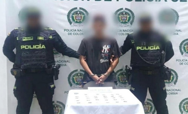 Dos funcionarios policiales con un ciudadano capturado frente a pendón institucional al frente una mesa sobre la cual están los elementos incautados