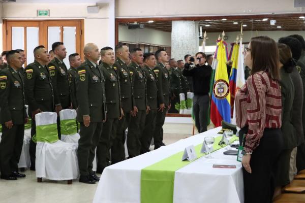 25 policías de pie acompañados de la mesa principal entonando el himno de la policía nacional, con las banderas de la policía nacional, Colombia y   Servicio Nacional de Aprendizaje SENA