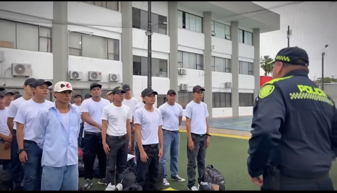 Se ve a un grupo de personas, la mayoría con camisetas blancas, reunidas en un espacio exterior frente a un edificio. Un oficial de policía se coloca al frente del grupo, de espaldas a la cámara, supervisando o dirigiendo el encuentro con las personas que buscan formar parte del cuerpo policial como auxiliares