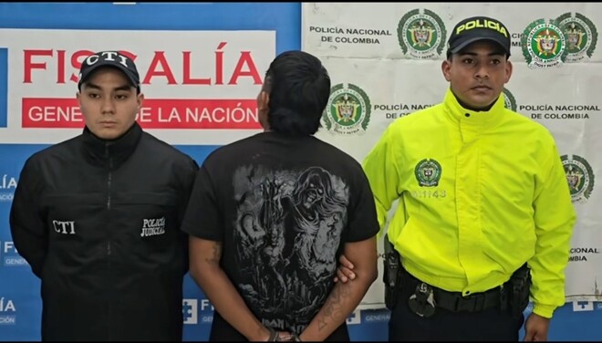 Un hombre esposado de espaldas, junto a un agente del CTI y un oficial de la Polic&iacute;a Nacional, ante el letrero de la Fiscal&iacute;a General de la Naci&oacute;n
