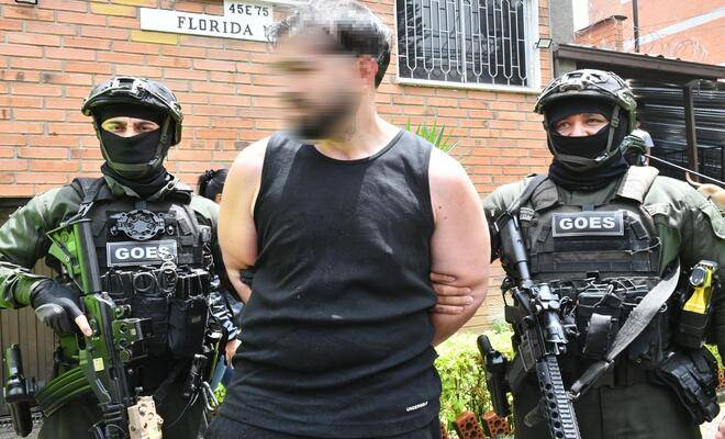 Exterior de edificio de ladrillo, en el centro hay una persona con camiseta negra y manos sujetas detrás, acompañada por dos personas uniformadas con equipo táctico que llevan chalecos con la palabra “GOES” y sostienen armas largas