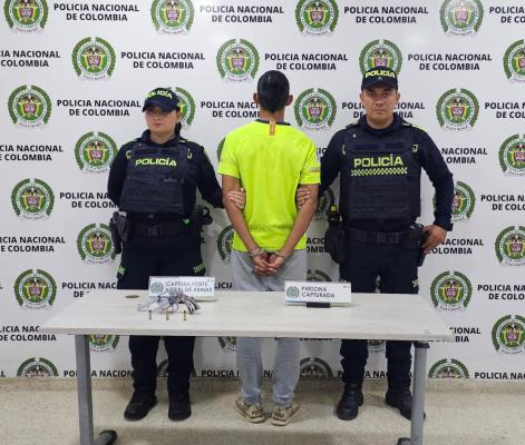 hombre capturado de camisa amarilla en medio de dos Polic&iacute;as, en frente una mesa blanca con arma de fuego incautada
