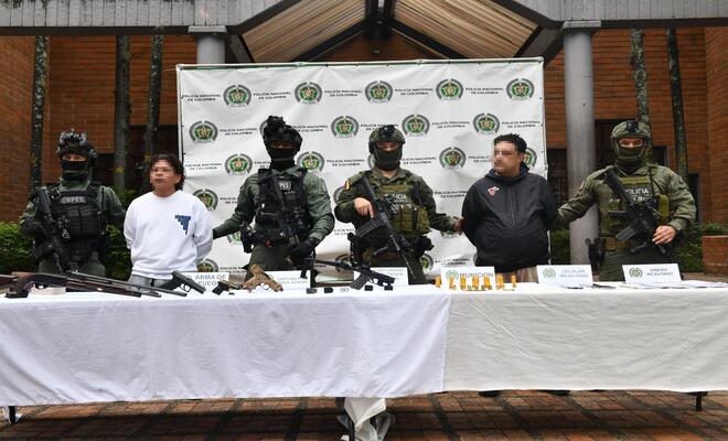 Varias personas de pie frente a un fondo institucional; en primer plano hay una mesa cubierta con mantel blanco donde se exhiben armas, municiones y otros objetos organizados, mientras personas uniformadas se ubican a los lados.