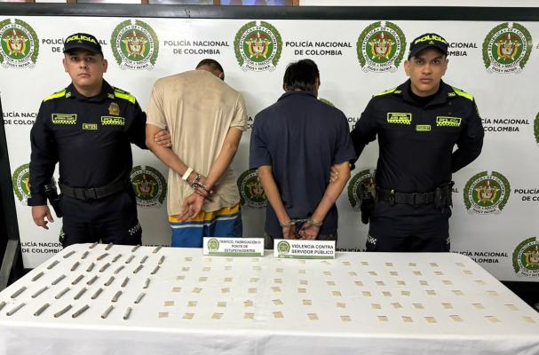 Dos hombres capturados en medio de dos policias, en frente una mesa con estuperfacientes incautados