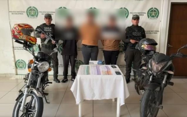 Hombres capturados en medio de dos policías, en frente dinero recuperado y a los lados motocicletas inmovilizadas