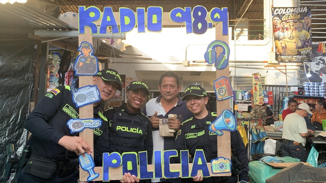 Agentes de la Policía comparten con un ciudadano en un mercado, utilizando un marco decorado con el logo de la emisora, promoviendo el acercamiento y la comunicación entre la institución y la comunidad