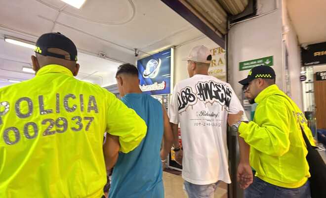 Pasillo interior de un centro comercial o edificio con locales; cuatro personas caminan juntas, dos de ellas con chaquetas reflectivas de la Polic&iacute;a Nacional de Colombia, mientras al fondo se observan avisos comerciales y se&ntilde;alizaci&oacute;n interior