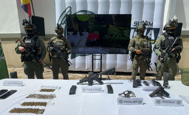 Hombres policías custodiando elementos incautados que se encuentran sobre una mesa con mantel blanco